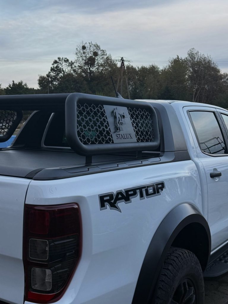 Tył białego samochodu typu pickup (model Ford Raptor) z zamontowaną czarną, stalową nadbudową skrzyni ładunkowej. W centralnej części ramy znajduje się metalowa płyta z wyciętym laserowo logo "STALUX".