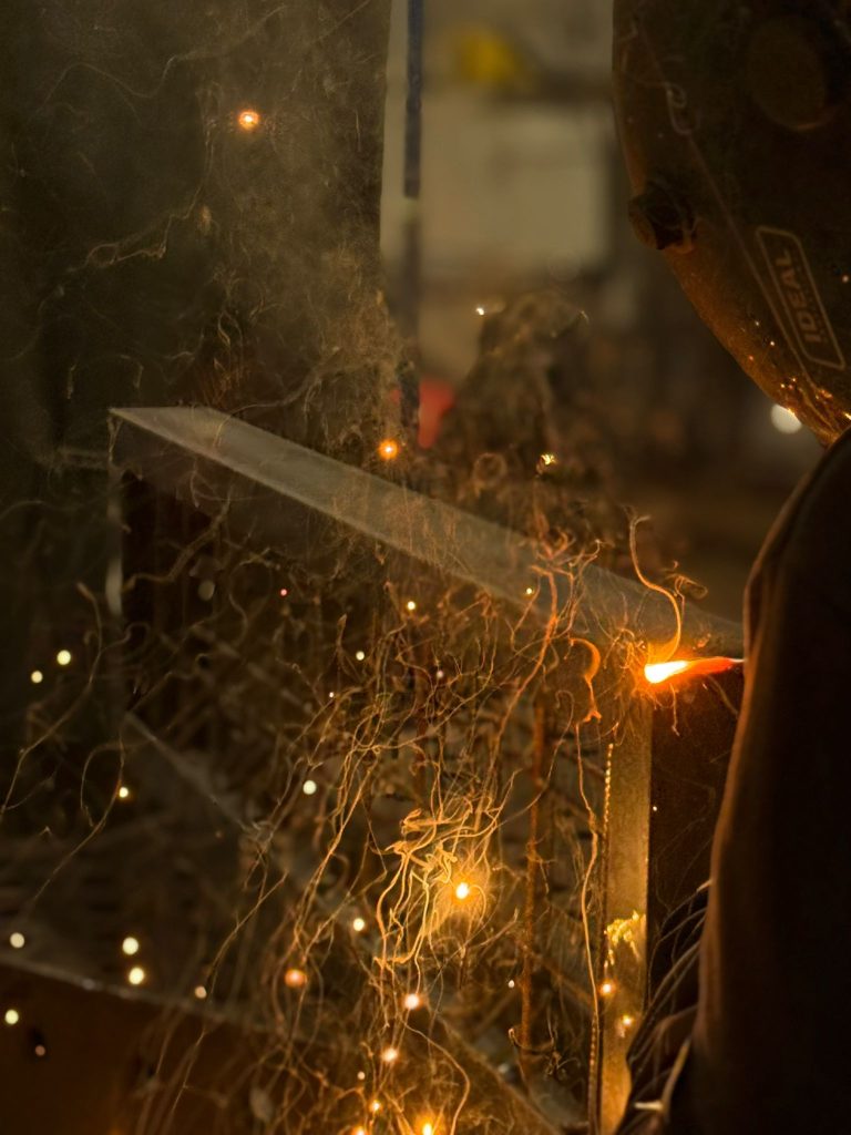 Zbliżenie na proces spawania elementów metalowych przez pracownika w masce ochronnej. Widoczne są intensywne, pomarańczowe iskry i rozbryzgi metalu na tle ciemnego warsztatu. Światło łuku spawalniczego oświetla konstrukcję.
